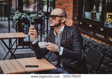 Pensive Modern Man Is Smoking A Cigarette Outside Of Coffeshop While Drinking His Cup Of Coffee.
