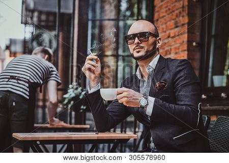 Pensive Modern Man Is Smoking A Cigarette Outside Of Coffeshop While Drinking His Cup Of Coffee.