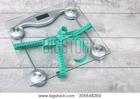 Green Tape Measure Bow Around Glass Weighing Scale On A Grey Rustic Surface. Healthy Weight Manageme