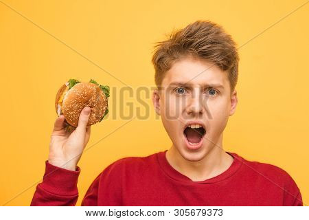 Portrait Of A Hungry Expressive Boy With A Burger In His Hand On A Yellow Background Looks Surprised