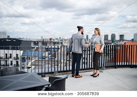Two Young Business People Standing On A Terrace Outside Office, Shaking Hands.