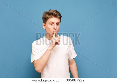 Guy In A White T-shirt Put His Finger To The Lips And Shows A Sign Of Silence, Isolated On A Blue Ba