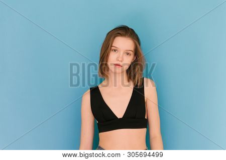 Attractive Girl With A Light Make-up Wears A Bra, Standing On A Blue Background, Looking Into The Ca