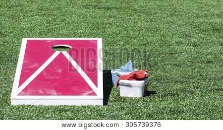 A Homemade Red And White Corn Hole Board On A Green Turf Field With Bean Bags In A Plastic Container