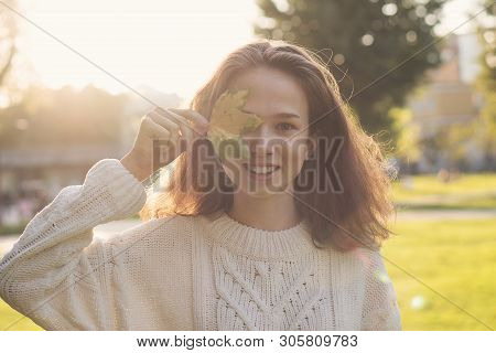Portrait Of European Charming Teen Girl. She Keeps Yellowed Fallen Maple Leaf. Model Poses In Rays O