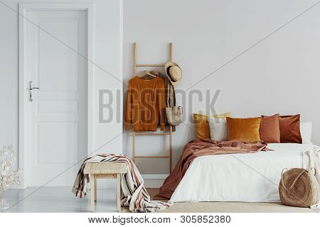 Sweaters, Wicker Hat And Bag On Wooden Ladder Next To King Size Bed With Velvet Pillows In White Bed