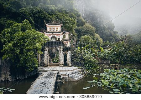 Scenery With Old Temple Against Karst Formation In Fog. Stone Bridge Over Lake Leading To Bich Dong 