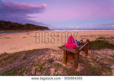 Woman Relaxes On Timber Bench Overlooking Beach In The Cool Hues Of Dusk