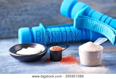 Creatine Powder, Scoop Of Whey Protein, Taurine Capsules And Blue Dumbbells In Background. Sport Nut