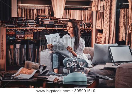Attractive Pensive Woman Is Sitting On The Sofa At Her Curtians Atelier While Handing Drawings About