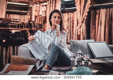 Attractive Pensive Woman Is Sitting On The Sofa At Her Curtians Atelier While Handing Drawings About