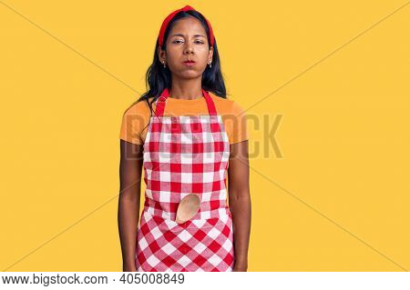 Young indian girl wearing professional baker apron puffing cheeks with funny face. mouth inflated with air, crazy expression. 