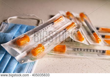 A Group Of New, Un-opened Medical Syringes In A Clinical Setting With A Protective Mask. Depicting T