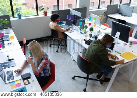 Diverse group of business people wearing face masks in creative office. group of people sitting at desks and using laptops. social distancing protection hygiene in workplace during covid 19 pandemic.