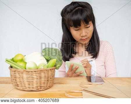 Woman Writing And Lood Mobile With Vegetables In Wicker Basket And Paper Bag On Wooden Table. Cook A