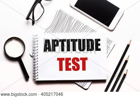 On A White Background Glasses, A Magnifier, Pencils, A Smartphone And A Notebook With The Text Aptit
