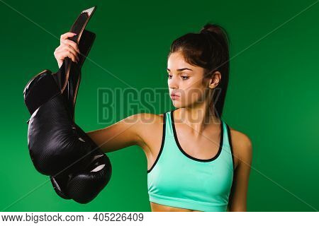 A Sporty Caucsian Brunette Girl Looks At The Black Boxing Gloves That She Holds In Her Hand On A Gre