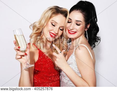 Two young girlfriends dressed in evening dresses, with gorgeous hairstyles are holding glasses of champagne and having fun. The blonde and the brunette are celebrating the New Year. Party time.