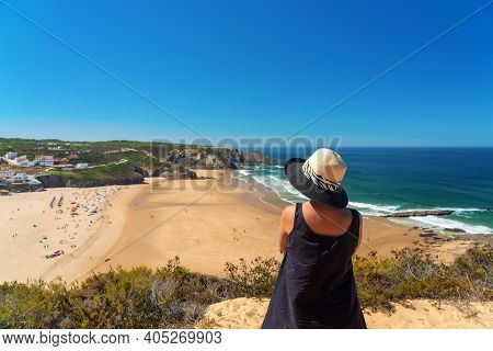 A Woman With A Hat On Her Head, On The Shore, Admires The View Of The Portuguese Beach, Odeceixe.