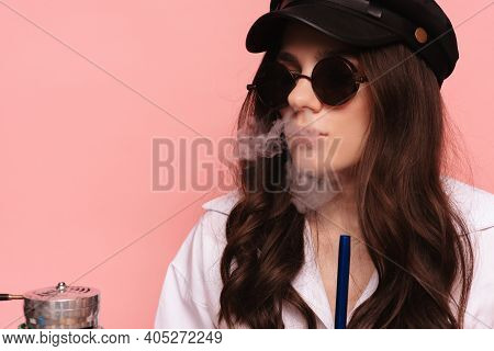 An Attractive Woman In A Cap And Glasses Smokes A Hookah, Sheesha And Enjoys Smoking. Pink Solid Col