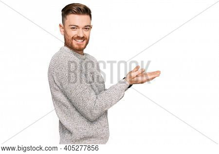 Young redhead man wearing casual winter sweater pointing aside with hands open palms showing copy space, presenting advertisement smiling excited happy 