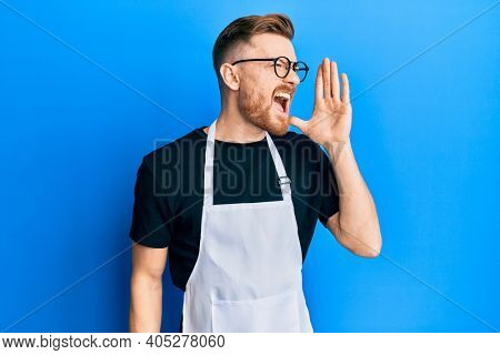 Young redhead man wearing professional apron shouting and screaming loud to side with hand on mouth. communication concept. 