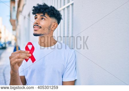 Young arab man smiling happy holding hiv awaraness red ribbon leaning on the wall.