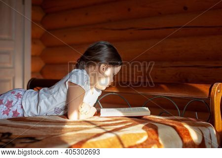Close-up An Enthusiastic, Interested Girl Lies On Her Stomach On A Blanket On The Bed And Reads A Bo