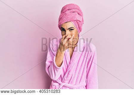 Young man wearing woman make up wearing shower towel on head and bathrobe smelling something stinky and disgusting, intolerable smell, holding breath with fingers on nose. bad smell 