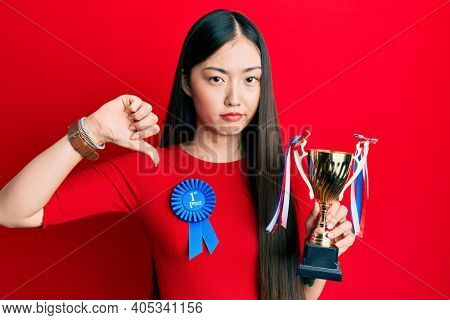 Young chinese woman wearing first place badge holding trophy with angry face, negative sign showing dislike with thumbs down, rejection concept 