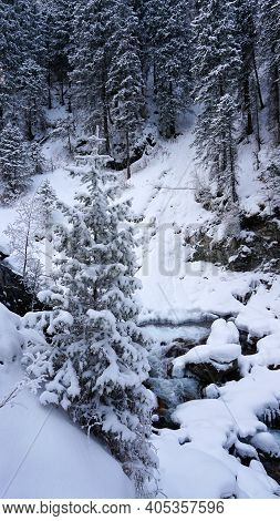A Clear Mountain River Runs Through A Snowy Gorge. There Is A Metal Bridge Covered With Snow. Tall S