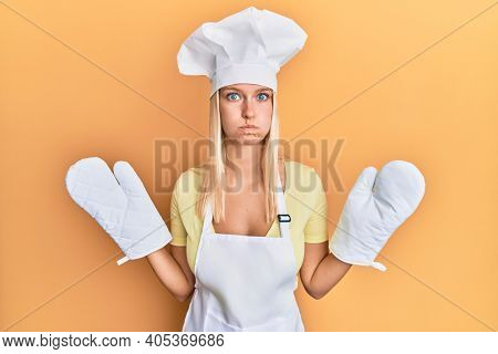Young blonde girl wearing baker uniform and cook hat puffing cheeks with funny face. mouth inflated with air, catching air. 