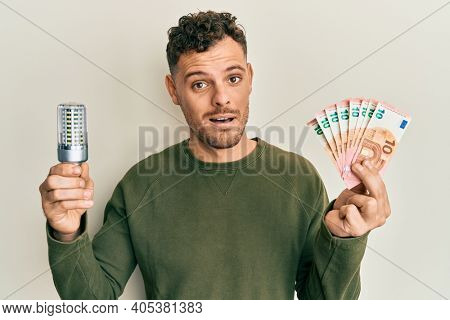 Young hispanic man holding led bulb and euros banknotes in shock face, looking skeptical and sarcastic, surprised with open mouth 
