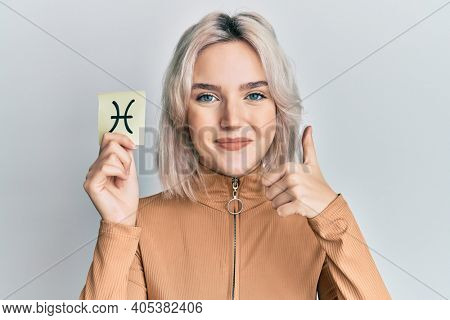 Young blonde girl holding paper with pisces zodiac sign smiling happy and positive, thumb up doing excellent and approval sign 