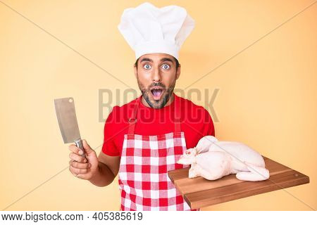 Young hispanic man cooking chicken holding knife afraid and shocked with surprise and amazed expression, fear and excited face. 