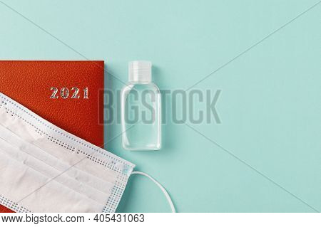 Medical Face Mask On A 2021 Year Red Leather Diary And Hand Sanitizer On A Pale Turquoise Background
