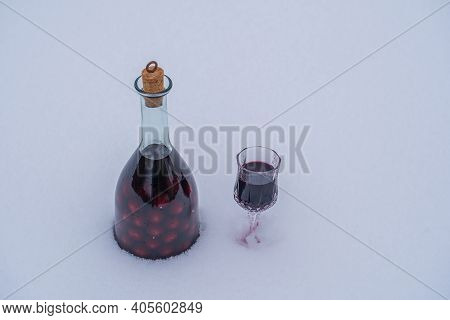 Homemade Tincture Of Red Cherry In A Glass Bottle And A Wine Crystal Glass On A Snow And White Backg
