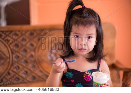 Little Child Girl Black-haired Sat And Scooped The Ice Cream From The Cup With A Blue Spoon. She Loo