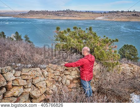 senior hiker at old stone fence overlooking frozen mountain lake  - Horsetooth Reservoir at foothills of Rocky Mountains - a popular recreational area in northern Colorado, fall or winter scenery