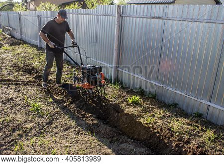 Male In The Spring Plows The Earth In Working Clothes With A Motoblock, Walk-behind Tractor