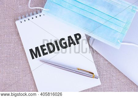 Medigap, The Text Is Written In A Notebook, Next To A Pen, A Disposable Medical Mask And A Laptop On