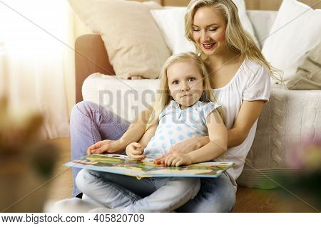 Happy Family. Blonde Young Mother Reading A Book To Her Cute Daughter While Sitting At Wooden Floor 