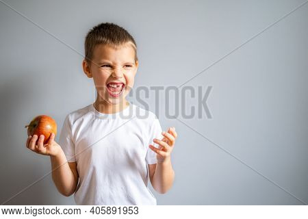 Boy Without Milk Upper Tooth In White T-shirt Holds An Apple In Hand On The Gray Background.