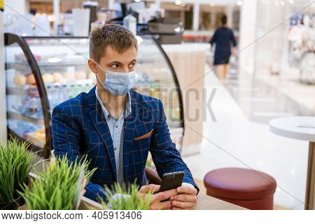 A Young Man In A Jacket Sits In A Cafe In A Medical Mask With A Phone In His Hand