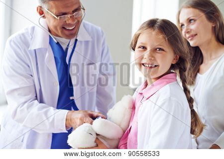 Adorable girl with teddybear looking at camera with her mother and doctor near by