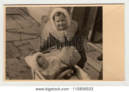 A vintage photo shows small children (girls) one of them is in a pram (baby carriage).