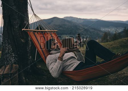 Leave all your worries behind. Handsome young man lying in hammock and reading a book while camping with his girlfriend
