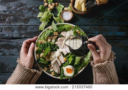 Female hands powring dressing to Classic Caesar salad with chicken breast in white ceramic plate. Served with ingredients above over old dark blue wooden background. Flat lay. Rustic style