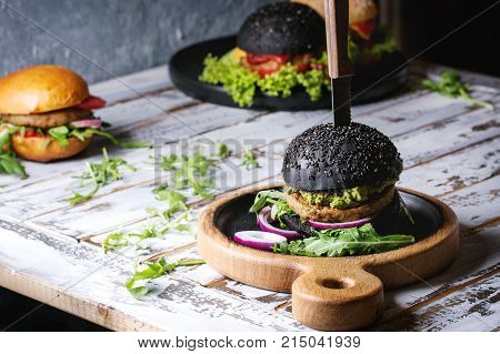 Set of homemade burgers in black and white buns with avocado, tomato sauce, lettuce, arugula, cheese, onion on wood serving board over white wooden plank table. Rustic style. Homemade fast food.