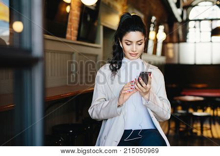 Pretty brunette woman with pony tail and appealing appearance sitting in cafe using her smartphone surfing internet using free wifi conncection and listening to her favourite music with earphones